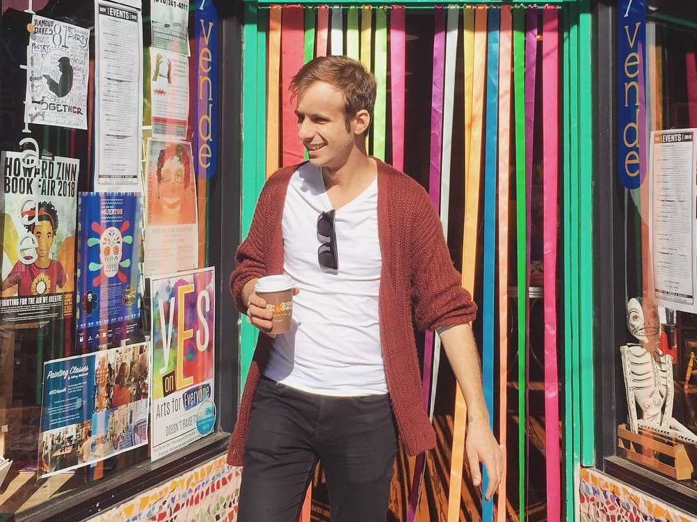 Photograph of Raphael Kabo standing outside Alley Cat Books in San Franscisco, smiling and holding a coffee. He's a young white man with light brown hair, wearing
a t-shirt, faded black jeans, and a burgundy knitted cardigan.